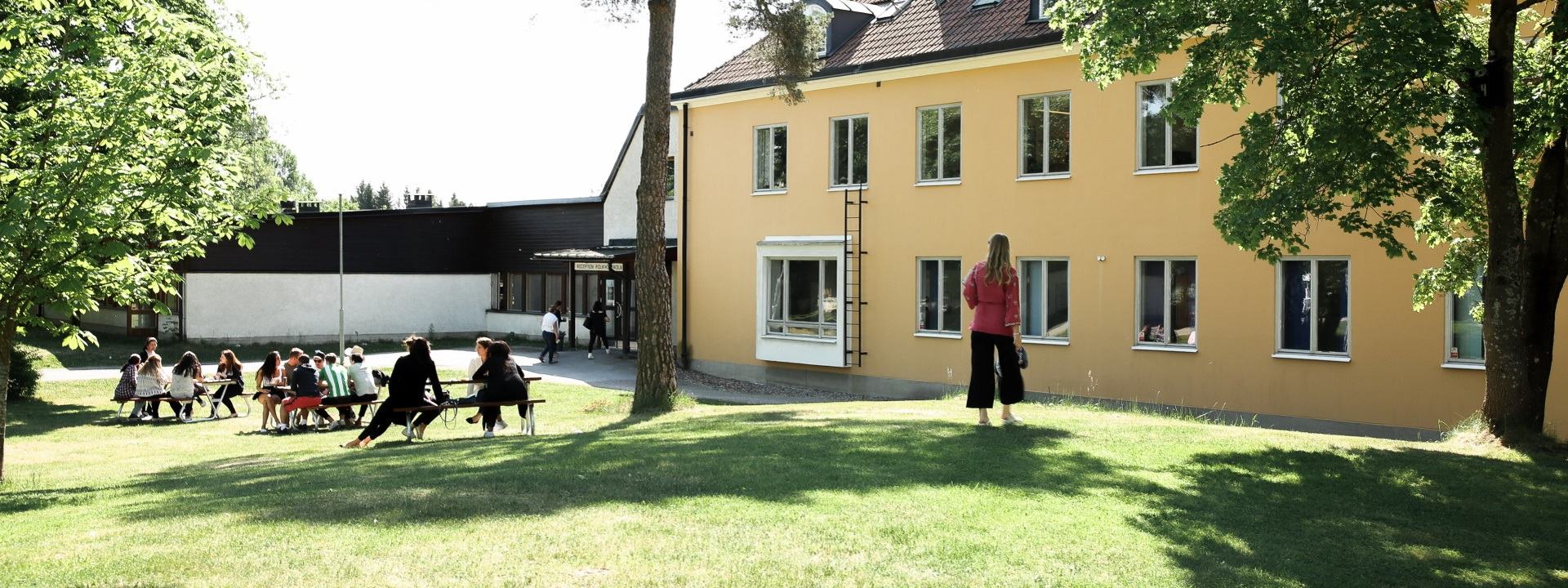 Studenter sitter och står på gräsmattan utanför en gul byggnad på Hagabergs folkhögskola.